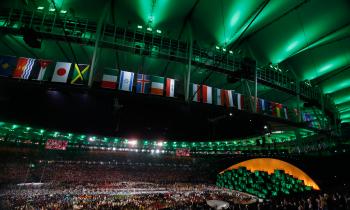 Foto do estádio do Maracanã durante a abertura das Olimpíadas do Rio, iluminado de verde escuro e completamente lotado, tanto no campo como nas arquibancadas. Na parte de cima da estrutura do estádio estão penduradas bandeiras de diversos países. No campo, estão diversos participantes da cerimônia de abertura. Em uma das extremidades do campo  localiza-se um painel com monitores verdes, adornado de uma espécie de cobertura amarela. Em cada monitor há a projeção de um rosto diferente. 