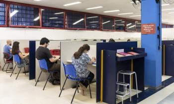 Foto de uma biblioteca. No centro da imagem estão quatro estudantes lendo um livro ou mexendo no computador. Eles estão sentados em cadeiras azuis. Na parte superior, há janelas vermelhas, algumas abertas, outras fechadas. No canto direito da imagem está uma espécie de armário azul escuro, com alguns livros sobre ele e uma pequena escada guardada abaixo. 