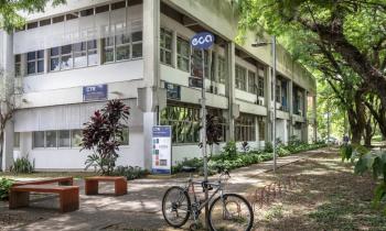 Foto do prédio do CTR. Em primeiro plano, há uma placa azul onde está escrito ECA em branco. No poste que sustenta a placa está amarrada uma bicicleta. Na parte esquerda existem três bancos laranja escuro, sem encosto. Mais ao fundo localiza-se o prédio, que foi fotografado de lado, de modo que é possível ver os dois lados dele. Na frente do prédio há placas em azul identificando o nome e a sigla do departamento. Ao seu lado encontra-se um caminho, ladeado de grama, arbustos e árvores. 