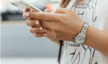 Foto de uma mulher segurando um celular. Não é possível ver o seu rosto, apenas da metade dos seus braços para frente. Em segundo plano, é possível identificar que ela veste uma camiseta branca estampada com dois elefantes. Suas unhas estão pintadas de branco e na na mão esquerda ela usa um relógio de pulseira prateada. 