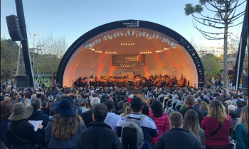 Foto de uma platéia assistindo a apresentação de uma orquestra sinfônica em um palco ao ar livre. Todos os assentos parecem estar ocupados, de modo que algumas pessoas ficam de pé no fundo. À frente do público está um palco com cobertura em meia-lua, no qual se posiciona a orquestra. A iluminação fica entre o laranja e o amarelo. No lado esquerdo do palco, encontra-se uma araucária. No direito, uma estrutura preta que sustenta o sistema de som. 