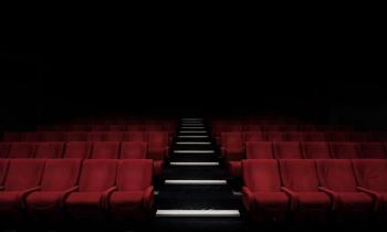 Foto de sala de cinema vazia. Ao centro, há um corredor de escadas. Várias fileiras de poltronas vermelhas podem ser vistas, todas elas vazias. O fundo da imagem é escuro, uma sombra.