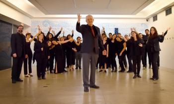 Foto dos integrantes do Coro de Câmara Comunicantus. Eles estão em uma sala. À frente do grupo, está regente e professor do coro. Ele é um homem mais velho, com cabelos curtos e brancos nas laterais da cabeça, usa um terno preto com gravata vermelha e está com uma das mãos levantadas. Ao fundo, está o grupo. Todos usam roupas pretas e olham para a câmera, com os braços levantados.