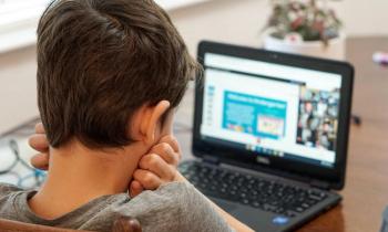 Foto de um menino sentado assistindo à aula online. Ele tem a pele branca, cabelos curtos e escuros, usa uma camiseta cinza e está com o rosto apoiado nas mãos semifechadas. O fundo é desfocado, mas é possível observar um computador sobre a mesa. O computador mostra uma apresentação de slides dividindo espaço com uma videochamada.