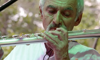 Foto de Toninho Carrasqueira, homem branco, idoso de cabelo brancos e bigode grisalho que usa uma camisa branca e toca uma flauta transversal. 