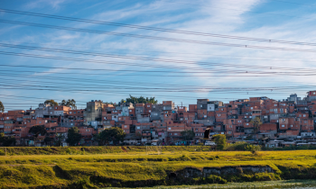 Foto de comunidade periférica próxima a um rio. Em primeiro plano, é possível observar parte do leito do rio e um campo de grama verde. Na parte superior, existem vários fios de alta tensão. Em segundo plano, há muitas casas próximas umas das outras, a maioria com tijolos e cimento aparentes. Existem poucas árvores no local. Ao fundo, um céu azul com algumas nuvens dispersas.