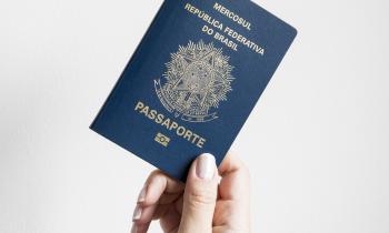 Foto. A frente de um fundo branco, uma mão, de uma pessoa branca, segura um passaporte, um livreto azul escuro com informações em dourado.