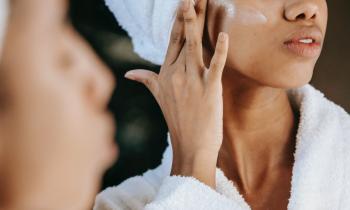 Foto de uma mulher negra com uma toalha no cabelo passando creme em frente ao espelho. No canto esquerdo da imagem, aparece o seu rosto desfocado. Em segundo plano e em foco, a mulher passa o creme em sua bochecha. É possível ver apenas seu nariz e sua boca. Ela veste um roupão de banho branco. 