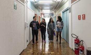 Foto de quatro estudantes andando em um corredor. À esquerda, um menino e uma menina estão conversando, eles são brancos e vestem roupas pretas. Ao lado deles, duas jovens se olham e sorriem, a da esquerda é branca e veste uma roupa preta com bolinhas pretas, a da direita é negra e veste cinza. No corredor, há algumas portas laranjas e azuis, as paredes são brancas.