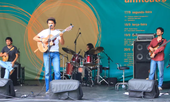 Um homem negro de cabelo escuro toca um violão e canta com um microfone de tripé. Ao fundo, um homem branco de cabelo escuro toca bateria. À esquerda, um homem amarelo segura um instrumento de cordas. À direita, um homem amarelo segura uma guitarra.