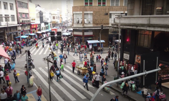 Print de um trecho do documentário 25 de Março: a singularidade dos fluxos e fixos. A imagem mostra, a partir de um ângulo elevado, as esquinas da rua 25 de março com a rua Cavalheiro Basílio Jafet e a Ladeira Porto Geral. Muitas pessoas estão nas calçadas e no meio da rua, vendendo ou comprando. 