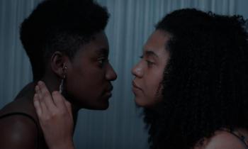 Foto de uma cena do curta. Duas mulheres negras se olham profundamente nos olhos. A da esquerda usa um brinco e tem cabelo crespo e curto. A da direita tem cabelo cacheado e volumoso. Ela coloca sua mão esquerda no pescoço da outra mulher. 