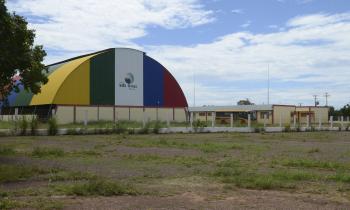 Foto da escola do quilombo Mata Cavalo tirada de longe. Em mais evidência, está a quadra da escola, que possui uma cobertura semicircular colorida. Mais ao fundo, está o prédio das salas de aula. O prédio é comprido e possui paredes amarelas, com listras vermelhas e muitas janelas. Na frente da construção, há um ônibus escolar amarelo. O chão é de terra e há nele muitas pedras, além de alguns pontos com grama.