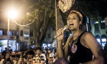 Foto de Marielle Franco, uma mulher negra que usa uma regata preta, brincos redondos pretos e uma faixa colorida no cabelo. Ela fala em um microfone. Há um adesivo lilás no seu peito e um livreto roxo na outra mão, ambos com seu nome escrito. Ao fundo, pessoas ouvem a vereadora. Entre essas pessoas, há dois fotógrafos, que apontam câmeras para ela.