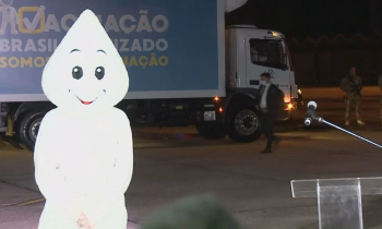 Foto do personagem Zé Gotinha sozinho na pista do aeroporto. Ele está ao lado de um pequeno púlpito de acrílico e ao fundo está estacionado um caminhão azul da campanha de vacinação. Na parte direita da foto, alguns soldados do exército armados se posicionam embaixo de um grande avião. 