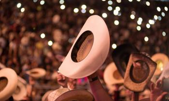 chapeu de cauboi é erguido por uma mãos de um homem branco, em meio a uma multidão de um show. ao redor, outros chapeus aparecem ergidos; ao fundo, luzes desfocadas