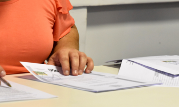 imagem de uma mesa sobre a qual estão alguns documentos; a mão de uma mulher branca, vestindo uma blusa laranja segura um desses documentos