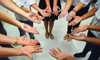 Foto dos braços e das palmas das mãos de várias pessoas. Elas parecem estar em uma sala fechada, com piso branco. É possível ver as pernas de alguns, mas o que se destaca é o gesto que todos fazem: estendem ambos os braços, com as mãos voltadas para cima, de modo a formar um círculo. 