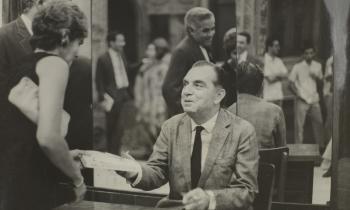 Foto em preto e branco. Ao centro, Nelson Rodrigues está sentado em uma cadeira, a frente de um espelho e atrás de uma mesa de madeira. É um homem branco de meia idade, de cabelo grisalho e usa um terno. Segura uma caneta com a mão esquerda e com a direita estende um livro a uma mulher, que usa um vestido preto, é branca e tem cabelo claro e curto. No reflexo do espelho ao fundo há uma fila de pessoas, algumas com um livro na mão.