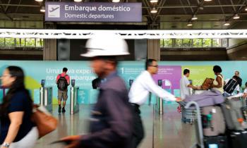 Foto do vai e vem de transeuntes com bagagens no terminal de passageiros do Galeão. Ao fundo, placa informativa  com o ícone de um avião subindo e os dizeres "Embarque doméstico" em português e inglês. 