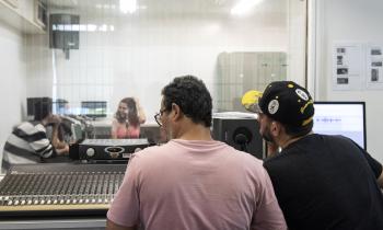 Foto de dois homens mexendo em uma mesa de mixagem de som. Eles estão de costas para a câmera. O da esquerda usa óculos e veste uma camiseta lilás, enquanto o da direita usa um boné e uma camiseta pretos. Ao fundo, por trás de uma tela de vidro, encontram-se duas mulheres e um homem gravando um material sonoro. À frente de cada um deles encontra-se um microfone.