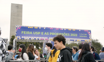 Foto de diversas pessoas caminhando entre as grades que organizam a fila de entrada para a Feira USP e As Profissões. Ao fundo, uma estrutura com o nome do evento e os dizeres "Boas Vindas". Atrás dessa estrutura está a Torre do Relógio.