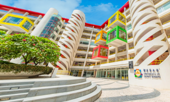 Foto da fachada da universidade. O prédio apresenta uma estrutura moderna, com três torres brancas espiraladas e alguns cubos coloridos projetados para fora da fachada. No canto inferior esquerdo da imagem, encontra-se uma parte elevada, na qual há um canteiro grande e florido.  