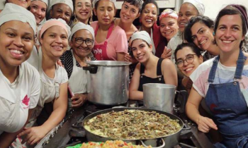 Foto de diversas mulheres ao redor de um fogão. Todas olham para a câmera e sorriem. A maioria usa toucas prendendo os cabelos. Em cima do fogão há grandes travessas com alimentos prontos e duas panelas.