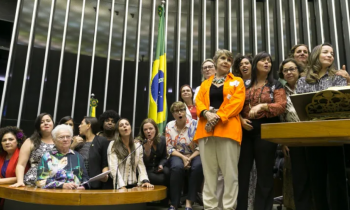 Foto de diversas mulheres, de diferentes idades e de maioria branca, na mesa e no púlpito da Câmara dos Deputados. Elas estão em pé, mas algumas estão mais altas do que outras devido ao desnível entre a mesa e o púlpito. Atrás das parlamentares está hasteada uma bandeira do Brasil.