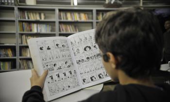 Foto de um menino branco, com cabelos curtos e castanhos que está sentado de costas. Ele lê um livro de histórias em quadrinhos. Ao fundo, estantes de livros.