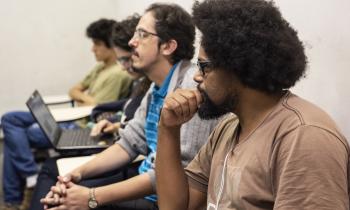 Foto de estudantes durante aula. Em primeiro plano, um homem negro de barba e cabelos escuros e crespos em penteado black power e óculos tem uma das mãos encostada no queixo. Atrás dele estão outros três jovens. Todos estão sentados em carteiras.