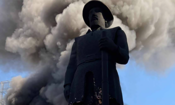 Foto da estátua do Borba Gato (de Santo Amaro), em chamas. A estátua está escura, coberta de cinzas. Logo atrás , aparece uma fumaça escura e densa. Há algumas chamas na parte inferior da imagem. Ao fundo,  o céu azul, sem nuvens.