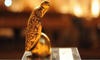 Foto do troféu do Prêmio Jabuti. É uma estatueta dourada de um jabuti apoiado sobre uma base arredondada. O jabuti é estilizado, sem patas, e seu casco é preenchido por entalhes das letras do alfabeto, de A a Z.