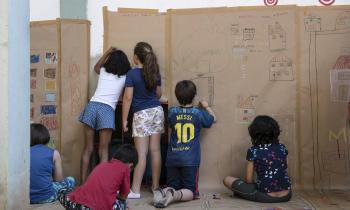 Foto de seis crianças desenhando e pintando em um mural de papel colado nas paredes e no chão. Algumas delas estão sentadas e outras estão em pé. 
