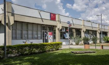 Foto da fachada do Departamento de Jornalismo e Editoração. Em primeiro plano, há um gramado ladeado por alguns arbustos. Mais ao fundo, está o prédio, onde encontram-se as placas do CJE e uma outra da ECA, que indica as direções para os outros departamentos. 