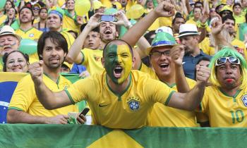 Foto de torcedores do Brasil em um estádio de futebol. Em primeiro plano, aparecem quatro torcedores, com destaque para um homem branco, vestido com a camisa da Seleção, que está com cada lado do rosto pintado de verde e amarelo e com os braços erguidos. Um outro torcedor usa um chapéu com as cores da bandeira, e um terceiro, uma peruca e óculos escuros. Todos os quatro olham para a câmera. 