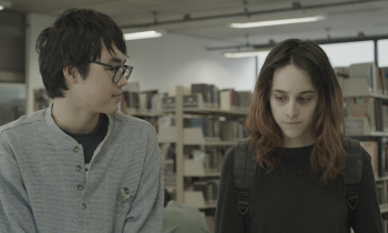 Imagem do filme Diário de Viagem. Um rapaz oriental de óculos observa menina de cabelo liso e pele pálida e com olhar baixo. Ao fundo uma prateleira de livros indicam que estão em uma biblioteca 