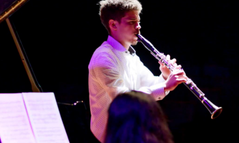 Foto de um homem tocando clarinete. Ele é branco, tem cabelos curtos e escuros e veste uma camisa branca. O fundo da imagem é totalmente preto, e o jovem é iluminado por luzes branca e roxa. Na porção inferior da foto, aparecem a cabeça de uma mulher que está de costas para a câmera e uma partitura apoiada no piano que ela toca.