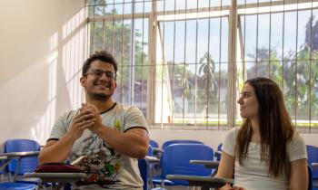 Foto de duas pessoas sentadas em uma sala de aula. Um homem sorridente está gesticulando com as mãos à esquerda. Ele é jovem, branco, tem óculos e cabelos escuros e veste uma camiseta clara com personagens desenhados. Uma mulher está sentada ao lado, ela olha para ele e sorri. A moça é jovem, é branca, tem longos cabelos lisos e escuros e veste uma camiseta branca. Atrás deles estão algumas carteiras escolares e uma janela.