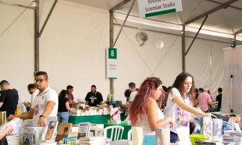 Foto do estande da editora Com-Arte na Festa do Livro da USP.  No expositor, os livros estão dispostos em uma mesa coberta por um tecido verde. Atrás dos livros, duas estudantes responsáveis pelo estade organizam as obras. Ao fundo, aparecem outros expositores do evento