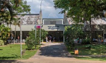 Foto da rampa de entrada para o prédio do Teatro Laboratório da ECA. As laterais da rampa têm um gramado arborizado. Ao fundo, bem no centro da imagem, a fachada do teatro. Há pessoas na entrada do prédio e cadeiras de plástico nas duas laterais da entrada. A luz do sol no chão indica que é um dia ensolarado.