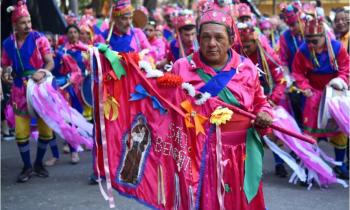 Foto de um senhor que veste um traje rosa de folia de reis e nas mãos usa uma bandeira com a imagem bordada de um santo e outros adornos. Ao fundo, foliões nas cores rosa e azul.
