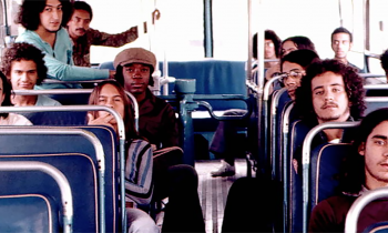 Foto de Milton Nascimento e do Clube da Esquina dentro de um ônibus antigo. São 13 homens ao total, 7 à direita e 6 à esquerda. Os bancos do ônibus são azuis e ele possui grandes janelas, que iluminam o interior do ambiente. 