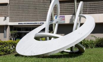 Foto de obra de arte sobre o gramado em frente à fachada do prédio principal da ECA. A escultura é branca, formada por 3 círculos e um semicírculo entrelaçados. Ao fundo, é possível ver a porta de entrada do prédio, com a inscrição Escola de Comunicações e Artes acima dela.