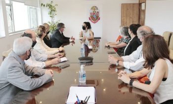 Foto de pessoas reunidas em uma grande mesa retangular. Na cabeceira da mesa, a cantora Marisa Monte - uma mulher branca, de cabelos escuros e ondulados, presos. Ela veste uma blusa branca e olha para a esquerda, onde está sentado o reitor da USP, um homem branco, de cabelos grisalhos e óculos. Ele veste um paletó cinza e uma camisa branca. Outras pessoas estão sentadas nos dois lados da mesa, homens e mulheres brancos. Ao fundo uma parede branca com o brasão da USP.