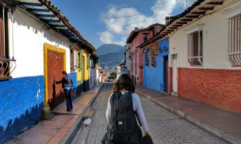 Foto de uma jovem que caminha sozinha por uma rua de Bogotá, na Colômbia. Ela está de costas para a câmera e carrega uma mochila preta nas costas. Seu cabelo é castanho e liso. Ela usa uma blusa cinza, de manga comprida, uma calça jeans e tem um casaco amarrado na cintura. A rua por onde ela passa é feita de tijolos e tanto do lado direito, quanto do lado esquerdo, existem casas em estilo colonial, coloridas nas cores branco, azul, rosa, verde, vermelho e amarelo.