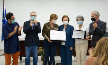 Foto de seis pessoas, três homens e três mulheres, em pé. Enquanto as mulheres seguram uma placa e um certificado, os homens aplaudem. Todos são brancos e usam máscara. Ao fundo, estão uma parede branca, uma mesa de madeira e uma bandeira azul hasteada. 