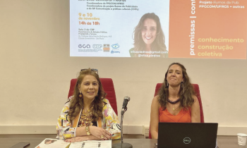 Foto de um seminário. Duas mulheres estão sentadas em uma mesa grande.  à esquerda, uma mulher branca, de cabelos longos e castanho claros. Ela tem um óculos apoiado sobre a testa. Veste uma blusa estampada e tem os antebraços apoiado um sobre o outro. à direita, uma mulher branca, de cabelos longos e castanho claros. Ela veste uma blusa laranja e um colar de madeira, marrom. à frente dela, um notebook aberto. Ao fundo, é projetada uma imagem da palestrante, título da palestra e outras informações.