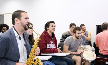 Foto de uma sala de aula. Mais a esquerda, o professor está sentado e tocando um saxofone, ao lado outros alunos estão sentados nas carteiras e olham para frente. Um deles segura um pandeiro e, outro, ao fundo, uma flauta.