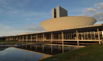 Foto do Congresso Nacional, mostrando em primeiro plano o espelho d'água, as janelas envidraçadas e a cúpula convexa da Câmara dos Deputados. Ao fundo, as torres centrais. 